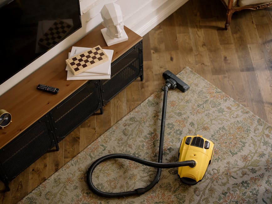 A person with curly hair, wearing a beige jacket and blue jeans, kneels on a patterned carpet in a room with warm lighting. They are opening a yellow and black upright vacuum cleaner, which is positio